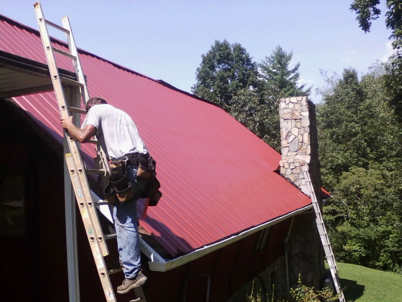 Roofer on a ladder beside a red metal roof and stone chimney — Soffit & Fascia Repair by Roofing Brothers in White Sulphur Springs